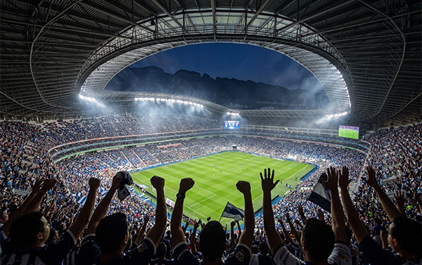 Aficionados con las manos arriba frente a la cancha y gradas de un estadio de fútbol lleno con las luces encendidas durante la noche